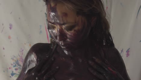 korean woman runs her hands across her face around and down her body, covered in a mix of paints while wearing a tank top in a studio against a white backdrop