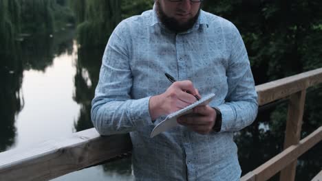 man makes notes in a notebook on the bridge by the river