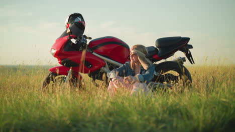 a woman sits next to her red bike in a grassy field, holding a piece of grass while gazing around thoughtfully, she wears a denim jacket, flower headband, and sunglasses