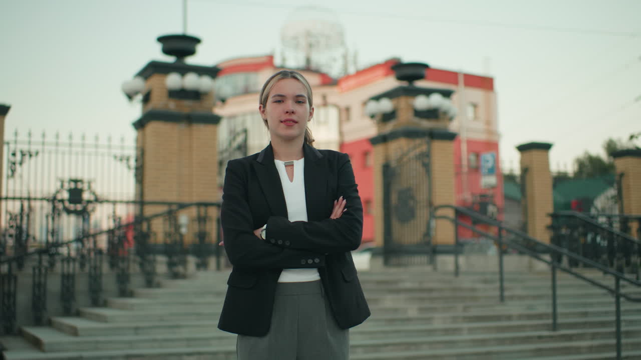 Pewna siebie biała kobieta stoi ze skrzyżowanymi rękami w scenerii miejskiej, ubrana profesjonalnie, emanuje śmiałością i opanowaniem, z elementami architektonicznymi i schodami w tle