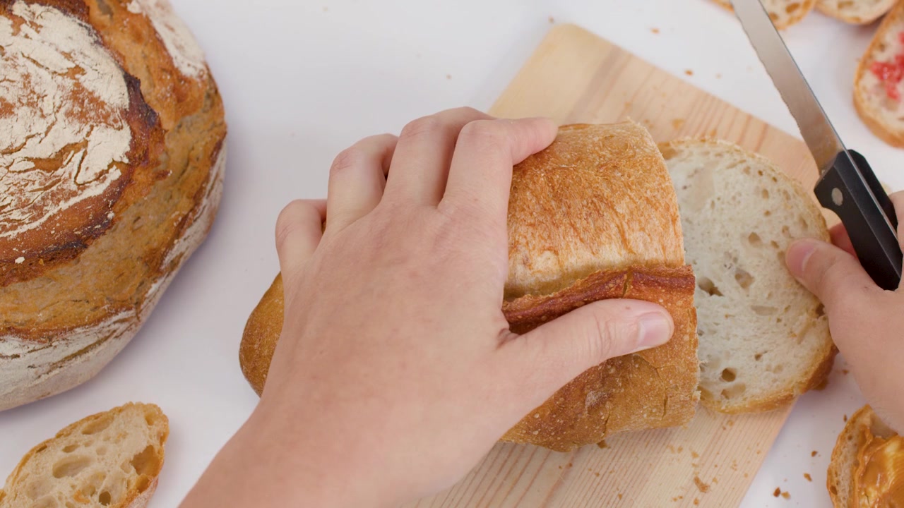 Top-down view of hands using a knife to slice crusty bread on a wooden board.