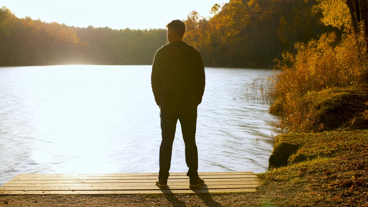 Una singola figura maschile sta su un molo di legno di fronte a un lago luminoso e retroilluminato, con le mani in tasca e i piedi alla larghezza delle spalle, indicando una quiete contemplativa, con dettagli di uomo, silhouette, molo e lago.