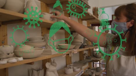 animation of virus icons over caucasian woman with face mask inspecting pottery