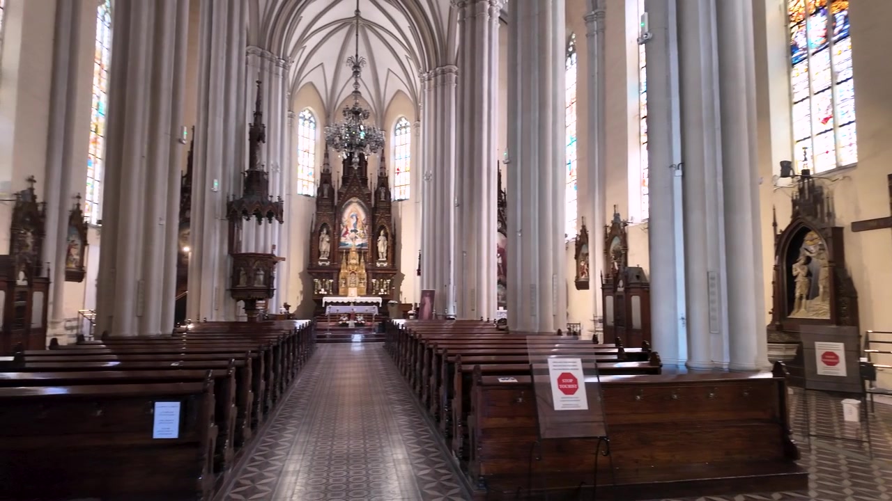 Le persone camminano attraverso una chiesa con alte colonne e luminose vetrate colorate. Lo spazio include panche di legno e statue religiose. Cartelli sono disposti per i visitatori.