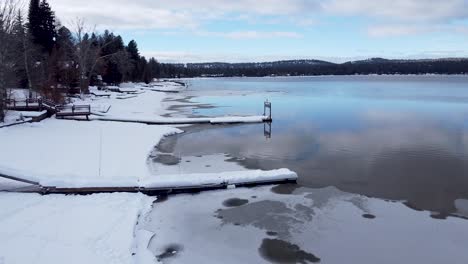 Snowy-icy-frozen-lake-in-winter-with-empty-snow-covered-boat-wharf-jetty-at-lake-front-houses-in-McCall,-Idaho,-USA---Aerial-drone-flyover