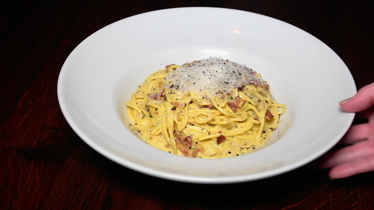 A close-up shot of a creamy pasta dish on a white plate, featuring spaghetti in a rich sauce with pancetta, topped with grated cheese and black pepper.