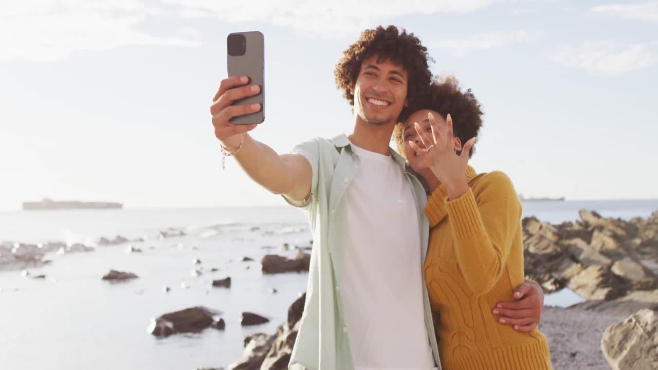 Coppia afroamericana che si scatta un selfie e mostra l'anello in piedi sulle rocce vicino al mare.
