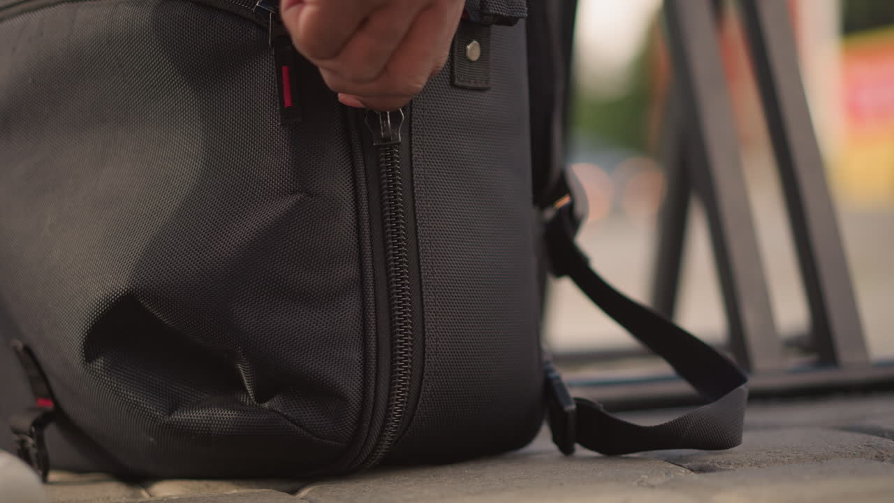 closeup torn backpack on pavement, black musician examines damaged dj bag near street setup, frayed zipper and ripped fabric, anxious hands kneeling, urban evening light, textured material, strap