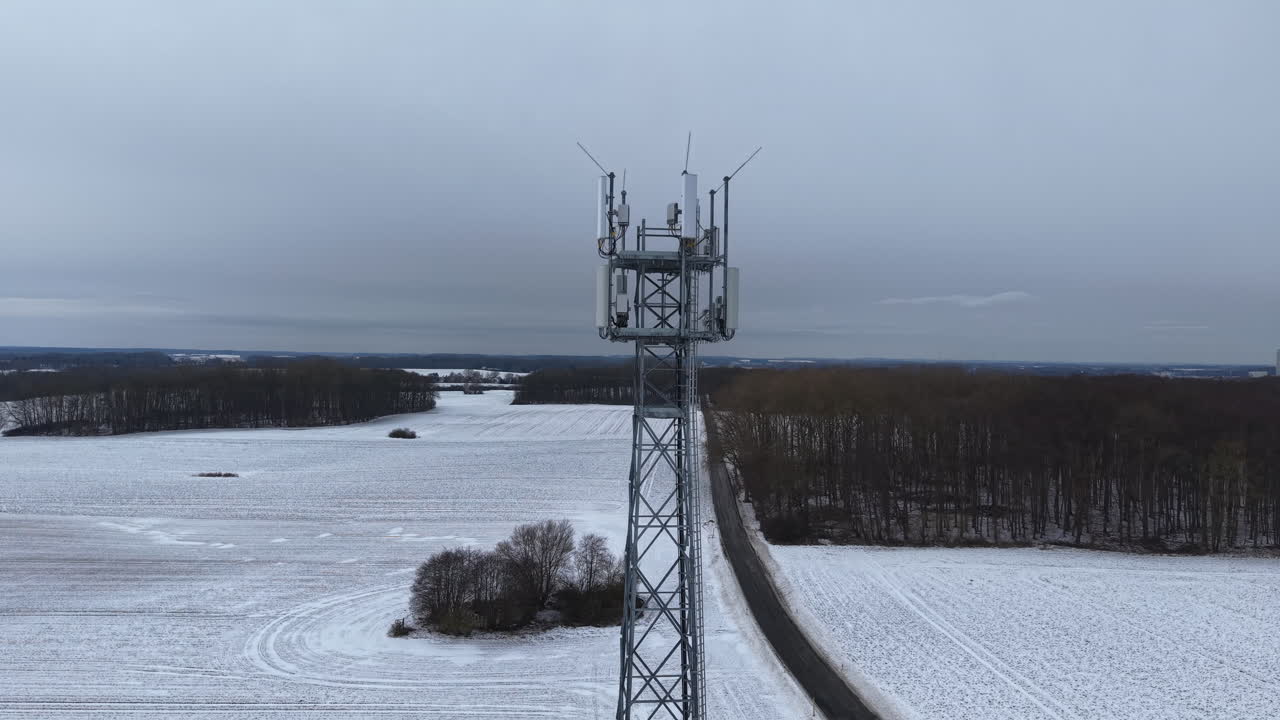 Aerial view of a modern mobile communication tower standing in a vast snow-covered winter landscape. The tall cellular mast with multiple antennas rises above frozen fields and distant forests