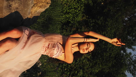 horizontal shot of full body portrait of gorgeous young brunette woman sitting on rocks and posing in exquisite pink ball gown surrounded by green vegetation