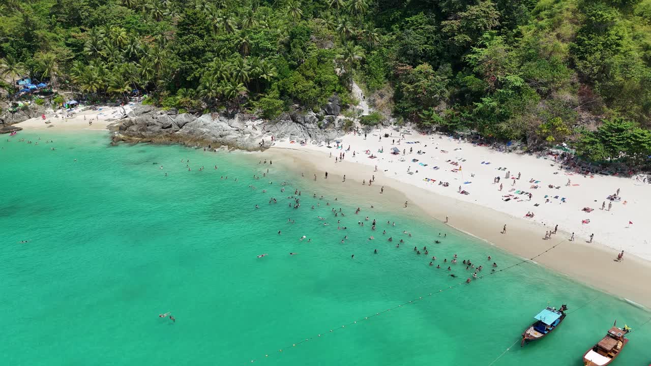 Drone footage captures a bustling Phuket beach with tourists swimming and relaxing under tropical sun, surrounded by lush greenery