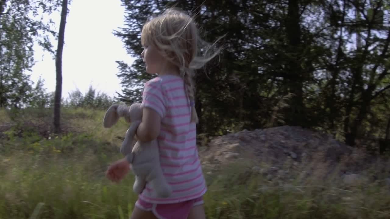 Elokuvamainen matala kuvakulma nuoresta ujosta tytöstä nallepehmolelun kanssa kävelemässä paljain jaloin kesällä, katsoen suoraan kameraan, suloinen viattomuus