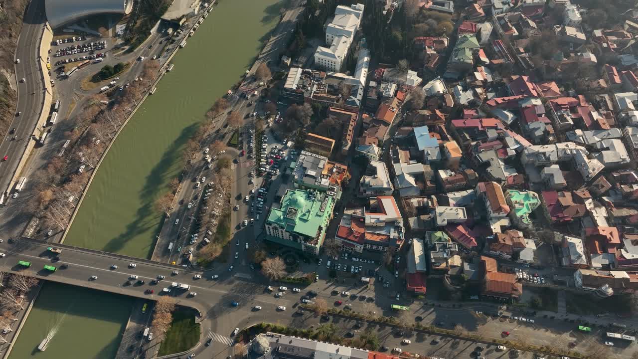 High-angle aerial photo showing two bridges over the Kura River and surrounding historic streets of Old Tbilisi. The shot combines water, roads, and colorful architecture in a busy city view