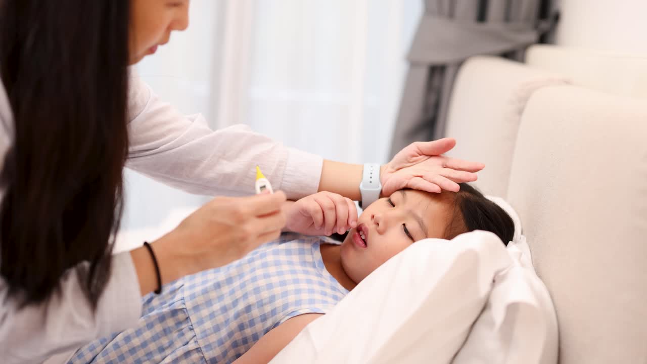 Concerned mother checks young daughter’s fever with thermometer in softly lit bedroom, close-up