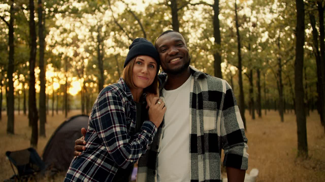 A happy couple, a guy with Black skin in a checkered shirt and a girl with a bob in a black hat, stand and smile and look at the camera during their hike in a sunny populated summer forest