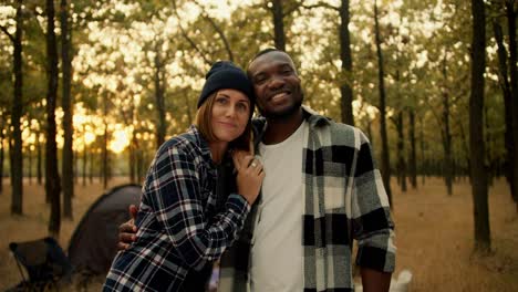 A-happy-couple,-a-guy-with-Black-skin-in-a-checkered-shirt-and-a-girl-with-a-bob-in-a-black-hat,-stand-and-smile-and-look-at-the-camera-during-their-hike-in-a-sunny-populated-summer-forest