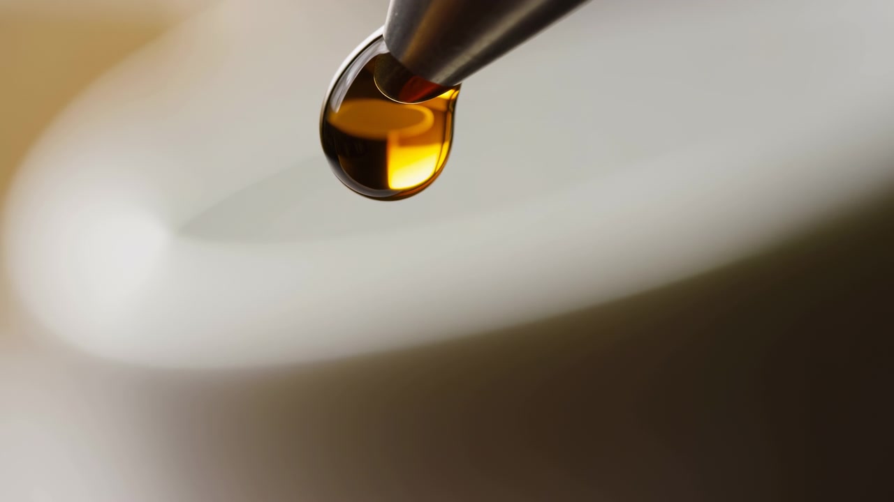 Gros plan d'un compte-gouttes métallique contenant un liquide ambré doré au-dessus d'une surface blanche lisse et incurvée, éclairé par une lumière douce et chaude et une faible profondeur de champ pour un rendu minimaliste et épuré