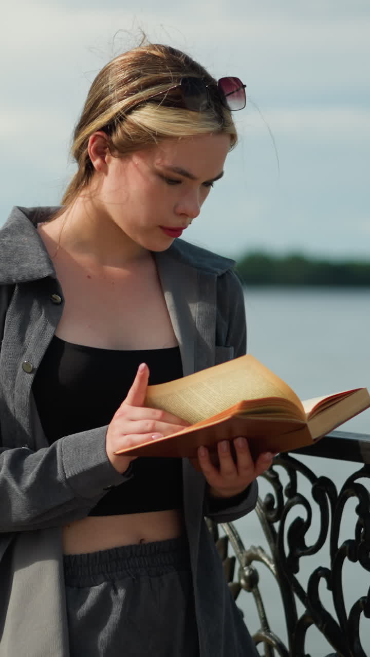 Young lady in grey clothing and black handbag stands by a fence, focused on reading a book as the wind blows the pages, she turns to a new page, with trees in the background and a lamp post nearby