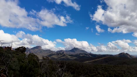 drone camera smoothly pans across rugged red rock cliffs, dense eucalyptus forest, and distant mountains under bright daylight and scattered clouds in coonabarabran, australia