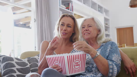 madre con hija adulta comiendo palomitas de maíz viendo una película en el sofá en casa juntos