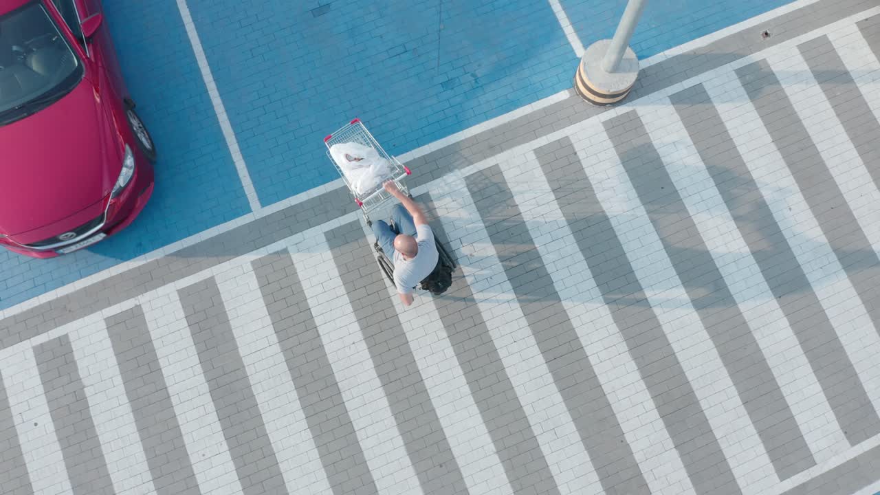 체어 에 앉은 장애인 이 슈퍼마켓 주차장 에서 자기 앞 에 바구니 를 밀고 있다