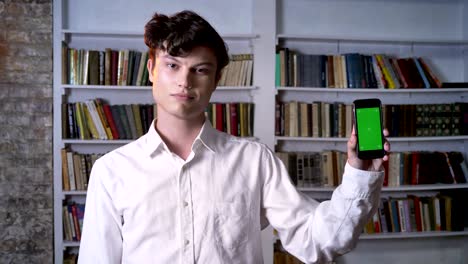 Brunette man is showing green screen of smartphone, watching at camera, communication concept, library on background