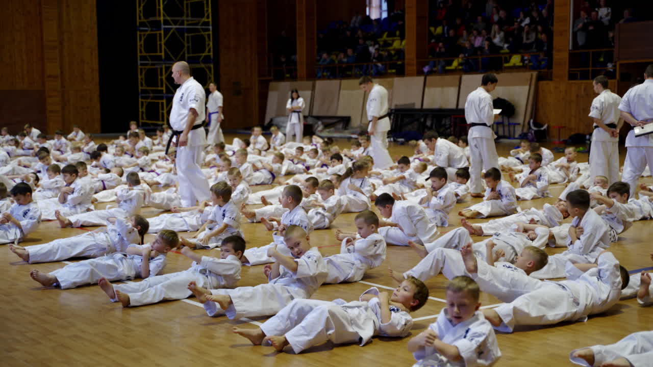 Young karate trainees in kimonos doing crunches in the gym. Coaches control the exercise performance. Audience sitting at backdrop.