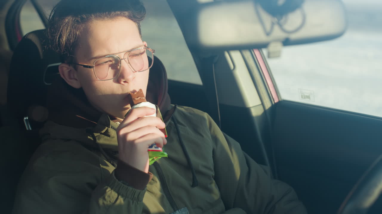 close up through windshield of boy biting into chocolate bar with sunlight reflecting on face and cozy hood framing head against blurred winter parking area backdrop