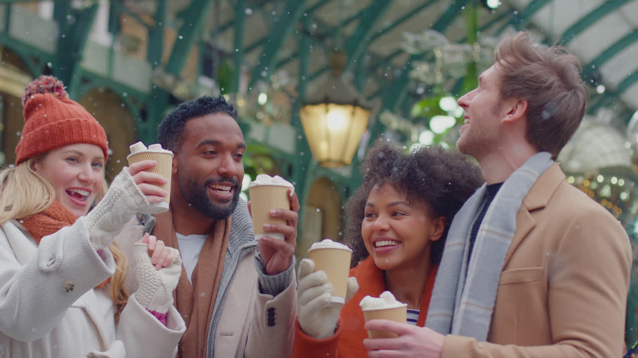 Group of friends drinking hot chocolate with marshmallows in snow at outdoor Christmas market in London's Covent Garden - shot in slow motion