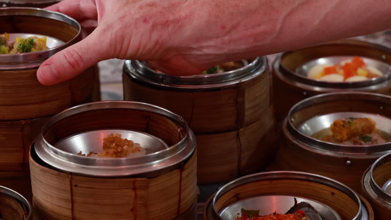 A hand selects dim sum from stacked bamboo steamers in a bustling Phuket setting, with warm lighting enhancing the vibrant atmosphere