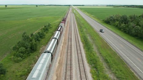 Vista-Aérea-Del-Viaje-En-Tren-De-Carga-A-Través-Del-Campo-Rural-Con-Carretera-Adyacente