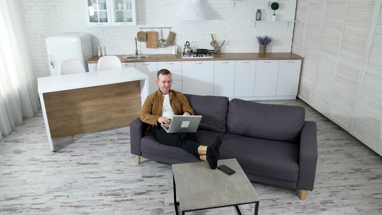 Young businessman working on a laptop. Modern kitchen background and man freelancer sitting in a couch with a laptop. Remote job.