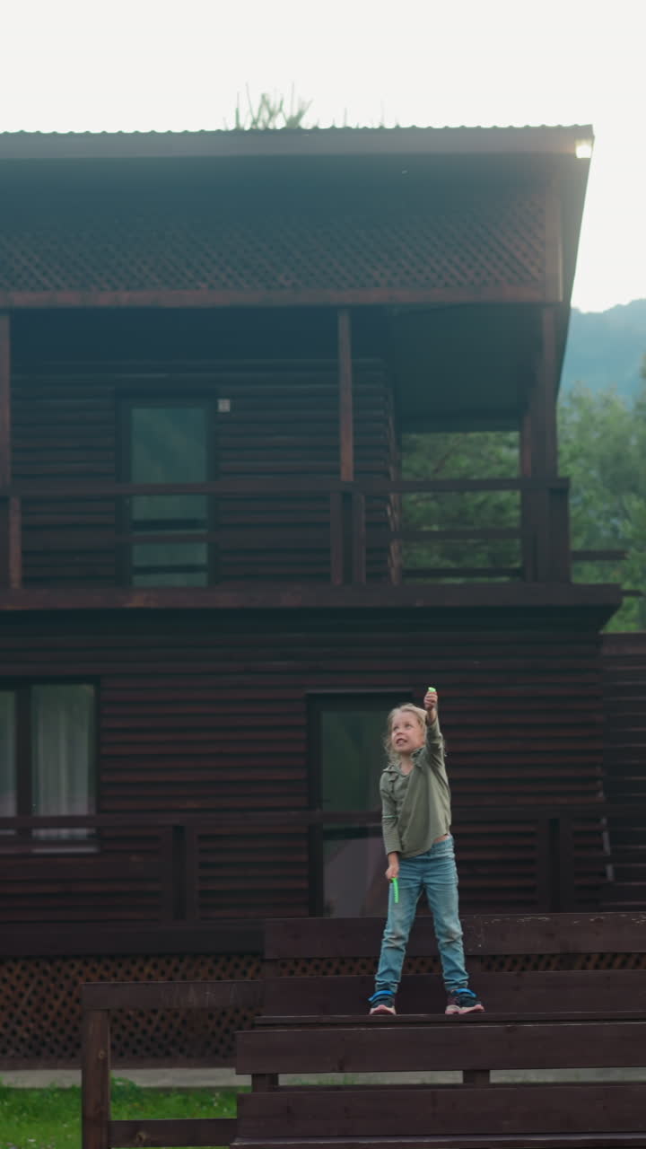 Little girl launches flying toy standing on wooden bench near eco hotel building in coniferous forest on gloomy spring day slow motion