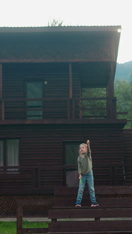 little girl launches flying toy standing on wooden bench near eco hotel building in coniferous forest on gloomy spring day slow motion