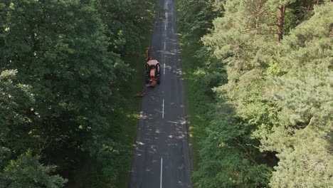 aerial view of a road maintenance tractor working along a narrow forest road. the scene is surrounded by dense green trees on both sides, capturing a peaceful natural environment with human activity