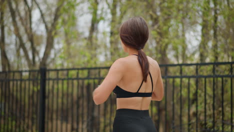 close-up view of young sporty woman stretching her waist outdoors, standing near iron fence with trees in background, enjoying fitness routine and healthy lifestyle, focusing on flexibility