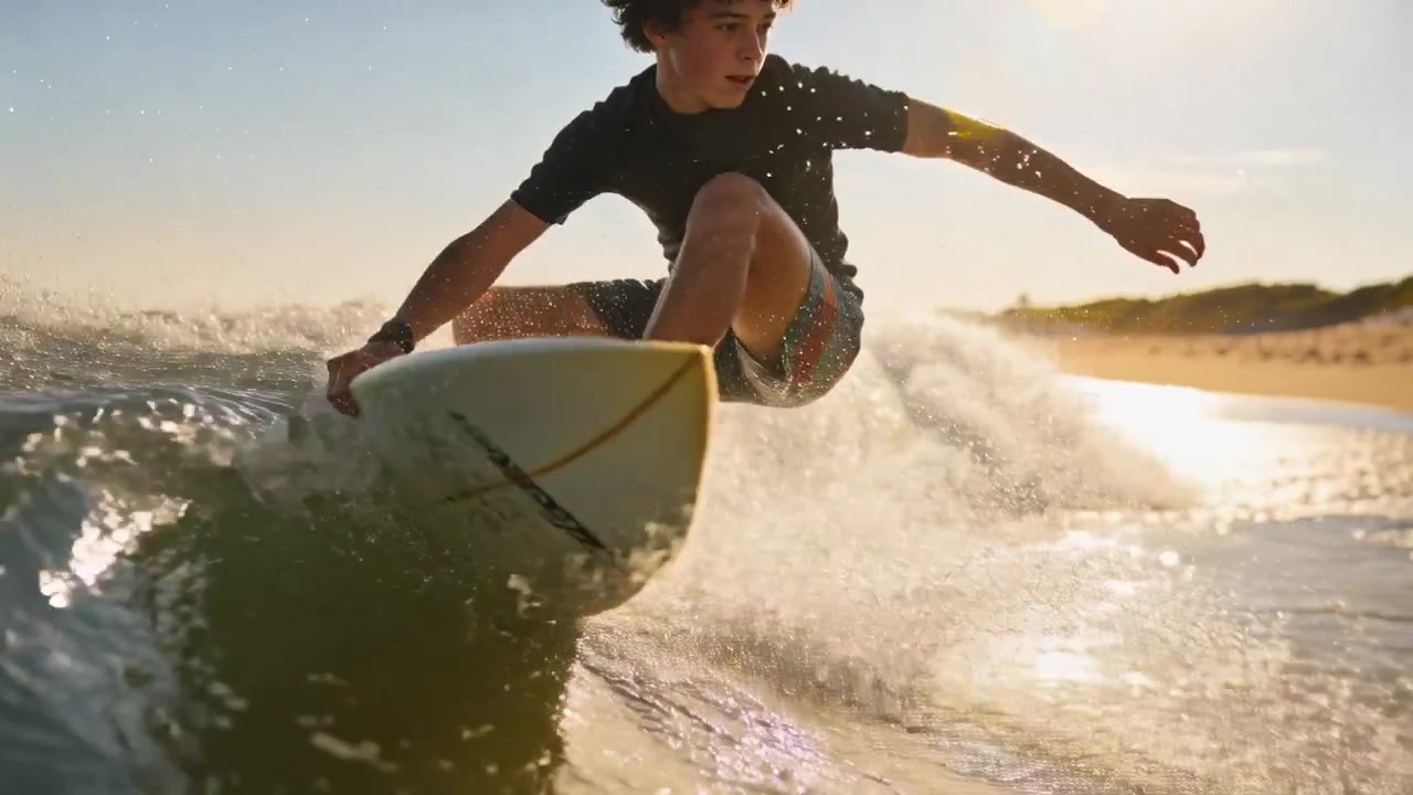 Miespuolinen surffaaja, yllään rashguard-paita, veistää aaltoa lyhyellä laudalla murtuvalla ranta-aallolla, etsien reittiä rannalle.