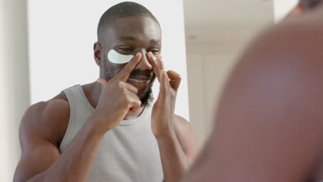 smiling african american man applying skincare patches under eyes at home