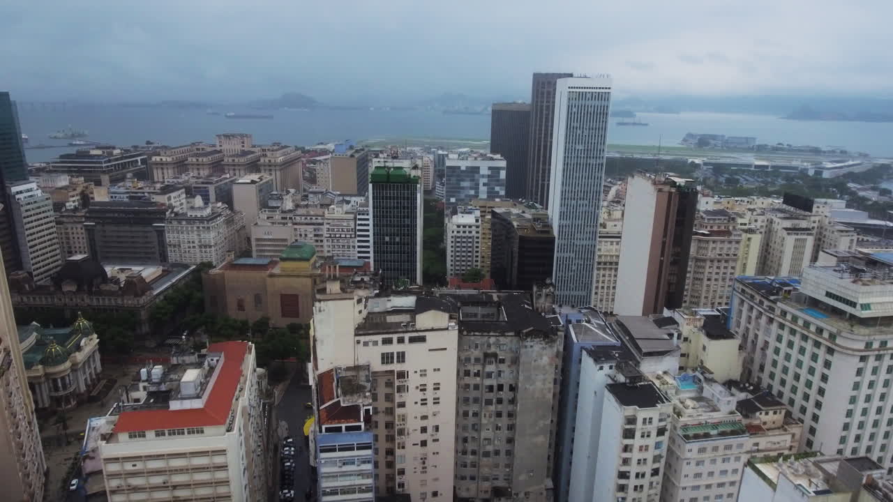 Imagem de drone recuando sobre o centro do Rio de Janeiro, mostrando densos edifícios altos, estruturas desgastadas e o oceano visível além do horizonte.