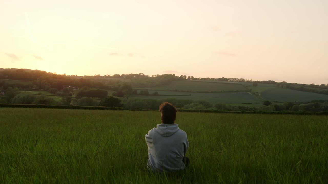 Rear view of a young man in jacket sitting in an idyllic countryside field gazing at sunset over lush horizon during sunset - static shot