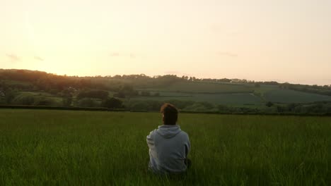 rear view of a young man in jacket sitting in an idyllic countryside field gazing at sunset over lush horizon during sunset - static shot