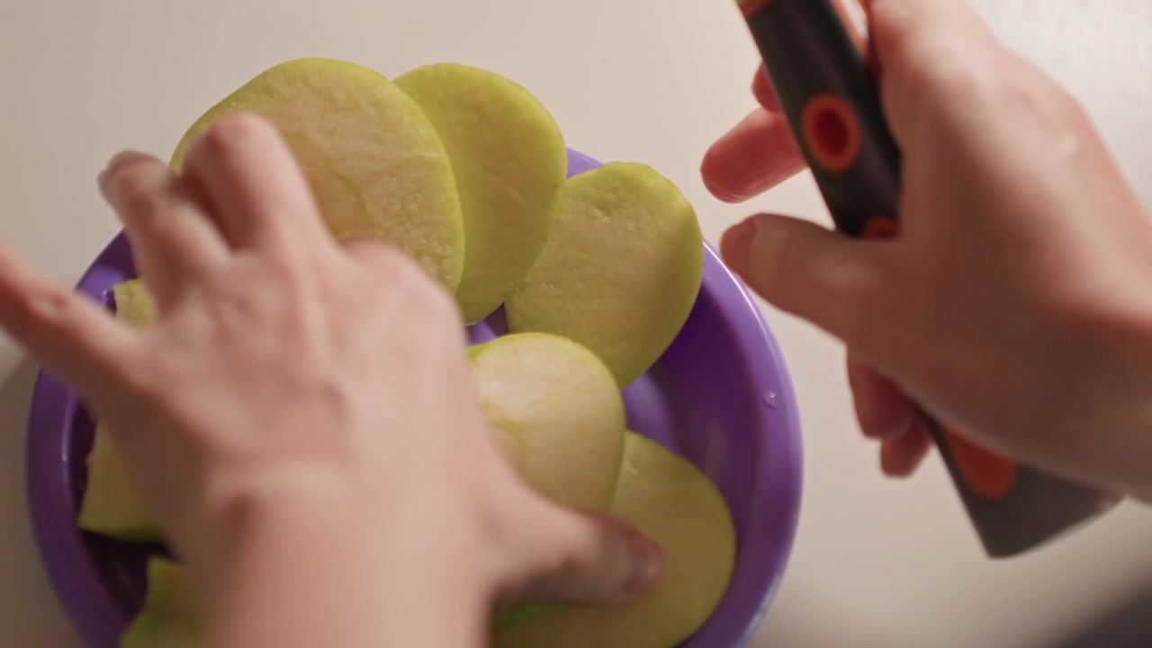 Primer plano de manos cortando manzanas verdes en rodajas finas. El proceso implica el uso de trabajo manual con cuchillo. El fondo liso mantiene el enfoque completamente en los movimientos de las manos con la manzana.