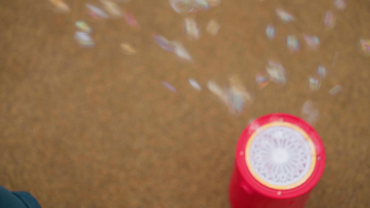 Top view close focus on red bubble machine on pavement releasing streams of soap bubbles upward, circular grille visible, soft blur background enhances shimmering motion