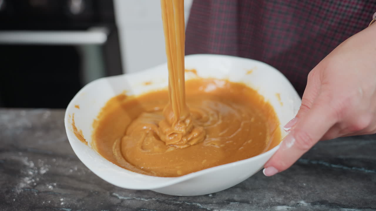 Baking process in action as whisk is lifted, causing thick batter to stretch and slowly drip into bowl, hands holding bowl and whisk are dusted with flour