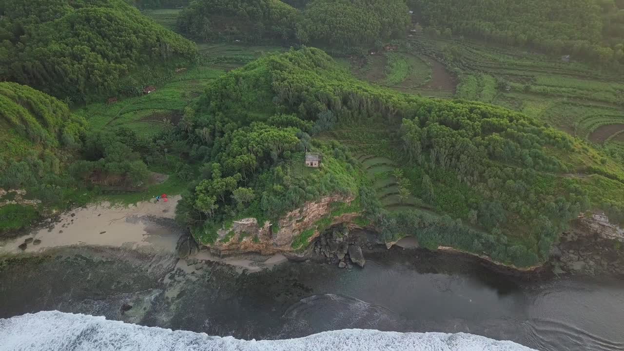Orbit drone shot of waves reaching green coastline of Indonesia with private house on cliff