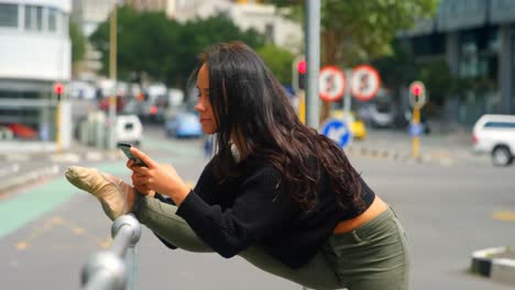 Bella-Mujer-Usando-Teléfono-Móvil-Mientras-La-Pierna-Estirada-Sobre-Una-Barandilla-En-La-Ciudad-4k