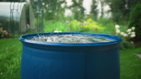 water drops into blue water drum, creating splashes as ripples form, garden background features blurred greenery, colorful flowers, and outdoor scene