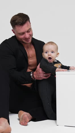 strong muscular man in black jacket on bare chest holds a baby. father and son studio footage. white backdrop. vertical video