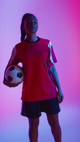 Vertical-Video-Studio-Portrait-Of-Female-Football-Or-Soccer-Player-Wearing-Team-Shirt-Turning-Around-Holding-Ball-Under-Her-Arm-Shot-Low-Key-Against-Colourful-Mixed-Lighting