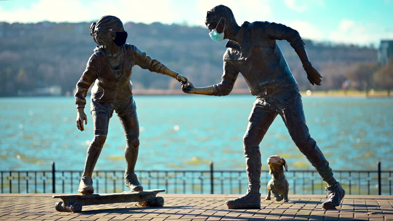 Bronze sculptures depict a child on a skateboard and an adult holding hands by the water. A dog stands nearby, capturing a joyful moment outdoors